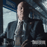a man in a suit and tie sits at a table with a cup of coffee in front of a sign that says daredevil born again