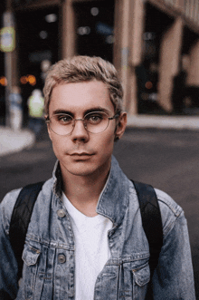 a young man wearing glasses and a denim jacket stands on a street