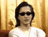 a man wearing sunglasses and a white shirt is sitting in front of a curtain .