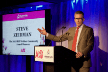 a man stands at a podium in front of a screen that says steve zeidman