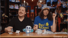 two men are sitting at a table with one wearing a shirt that says buy and leave