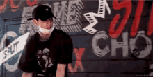 a man wearing a baseball cap and a mask is standing in front of a wall with graffiti on it .