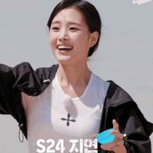 a woman wearing a white shirt and a black jacket is smiling and holding a blue container with the letters s24 on it