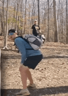 a man with a backpack is squatting on a dirt path in the woods .