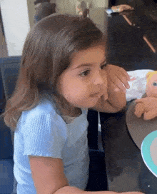a little girl is sitting at a table with a doll and eating a spoonful of food .