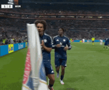 two soccer players are running on a field with a scoreboard that says mun