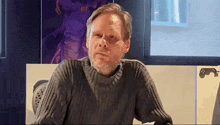 a man with a beard is sitting at a table wearing a sweater and looking at the camera .