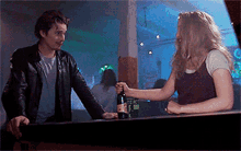a man in a leather jacket holds a bottle of beer while a woman looks on
