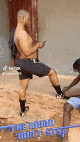 a man without a shirt is standing on one leg while another man ties his shoe .