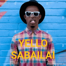 a man wearing sunglasses and a plaid shirt stands in front of a blue brick wall that says yello sabaila
