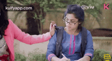 a girl with glasses and a backpack is sitting at a table with a pen in her hand while another girl touches her shoulder