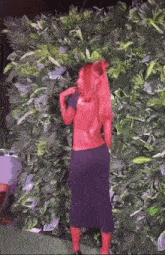 a woman with red hair and a black dress is standing in front of a wall of plants .
