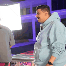 a man in a light blue hoodie is laughing while standing next to a man in a grey shirt .