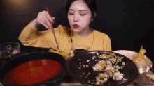 a woman in a yellow sweater is eating a plate of food with chopsticks