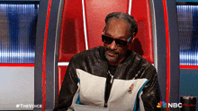 snoop dogg is sitting in a chair with a notebook in front of him and a nbc logo in the background
