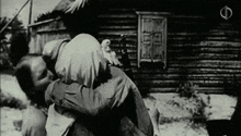 a black and white photo of a man carrying a woman in front of a log cabin