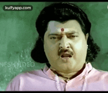 a man with a mustache and a red dot on his forehead is wearing a pink shirt .