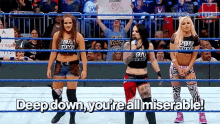 three women are standing in a wrestling ring with the words `` deep down you 're all miserable '' written on the bottom .