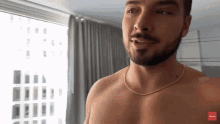 a shirtless man with a beard and a gold chain around his neck is standing in front of a window