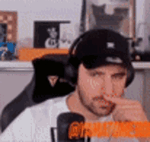 a man wearing headphones and a hat is sitting in front of a microphone and holding his nose .