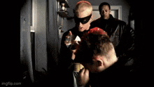 a man in a mask is holding a red object in front of a man in a black jacket .