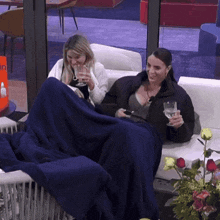 two women are sitting on a couch with a blue blanket and drinking wine