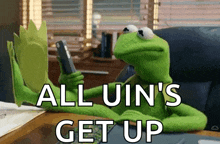 kermit the frog is sitting at a desk holding a cell phone and says all uin 's get up .
