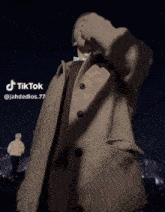 a man in a tan coat is standing in front of a city at night and has a tiktok account