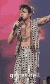 a shirtless man is singing into a microphone while wearing sequined pants and a vest .