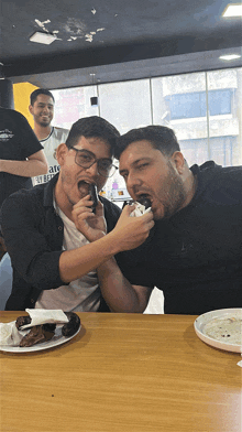a man wearing a shirt that says " fly better " is eating a piece of food with another man