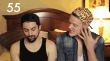 a man wearing a leopard print hat talks to another man in front of a bed with the number 55 on it