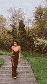 a woman in a long brown dress is walking down a wooden path holding a glass of wine