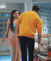 a man and a woman are standing next to each other in a living room . the man is wearing a yellow sweater .