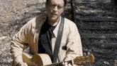 a man in a tan jacket is holding a guitar