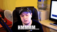 a man wearing a hat is sitting in front of a computer with the words hmmmm written on his face
