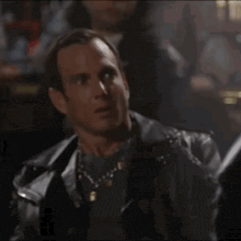 a man wearing a black leather jacket and a necklace is sitting in a dark room .