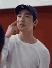 a young man wearing a baseball cap and a white sweater is pointing at his face .