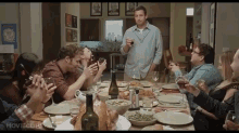 a group of people sitting around a table with a man standing in front of them with a glass of wine in his hand