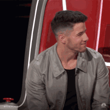 a man in a gray jacket is sitting in a red chair on the voice show