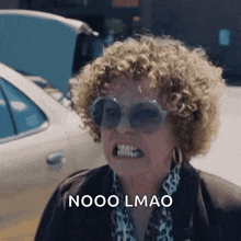 a woman with curly hair and sunglasses is making a funny face while standing in front of a car .