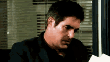 a man in a black suit is sitting at a desk reading a piece of paper .