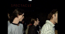 a man in a gray shirt is standing in a crowd with the word spectacle on the bottom right
