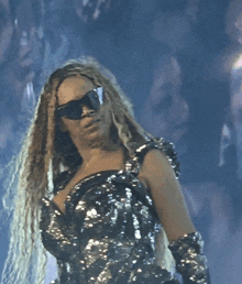 a woman wearing sunglasses and a sequined dress stands on a stage