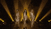 three women in gold dresses are dancing on a stage with yellow lights behind them