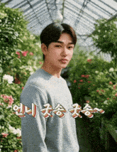 a young man in a blue sweater stands in front of a greenhouse with flowers