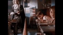a group of children are sitting at desks in a classroom while a man in a costume stands behind them .