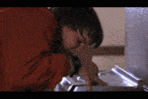 a young boy in a red shirt is drinking from a water fountain