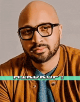 a bald man with glasses and a beard is wearing a brown jacket and a blue shirt