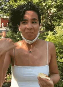 a woman wearing a white tank top and a face mask holds a piece of food