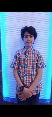 a young boy in a plaid shirt and tie is standing in front of a blue wall with his hands folded .